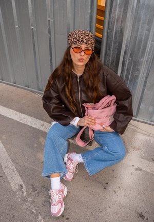 Joven con chaqueta de cuero marrón, pañuelo de leopardo en la cabeza, gafas de sol naranjas, vaqueros azules y zapatillas rosas sentada en el pavimento sosteniendo una bolsa rosa.