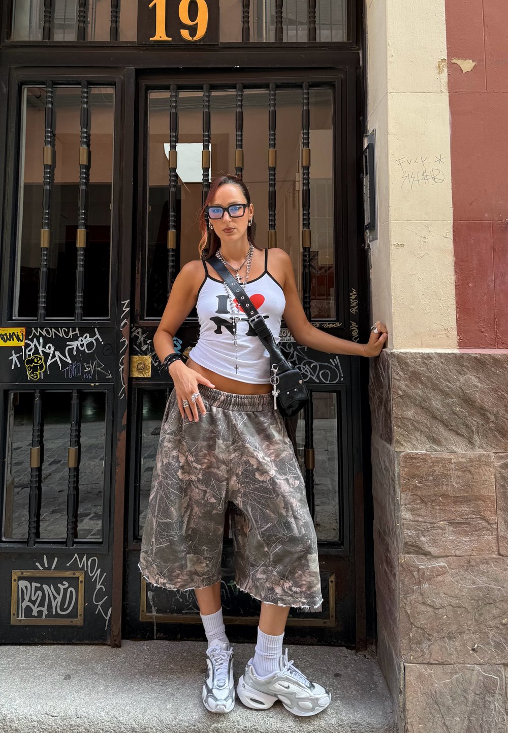 A woman wearing an I ❤️ NY tank top, oversized camo shorts, white sneakers, sunglasses, and necklaces is standing in front of a black barred door marked with the number 19.