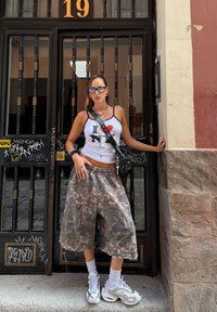 A woman wearing an I ❤️ NY tank top, oversized camo shorts, white sneakers, sunglasses, and necklaces is standing in front of a black barred door marked with the number 19.