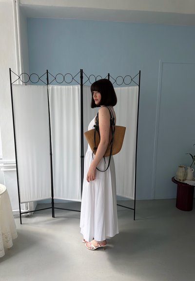 Mujer con vestido blanco y sandalias está de pie en el interior sosteniendo un bolso tote marrón, con una pantalla plegable blanca y una pared azul claro detrás de ella.