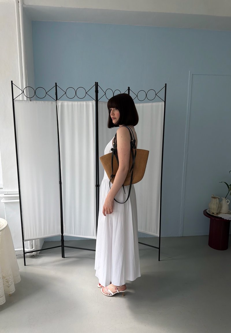 Vrouw in witte jurk en sandalen staat binnen en houdt een bruine draagtas vast, met een wit klap-scherm en een lichtblauwe muur achter haar.