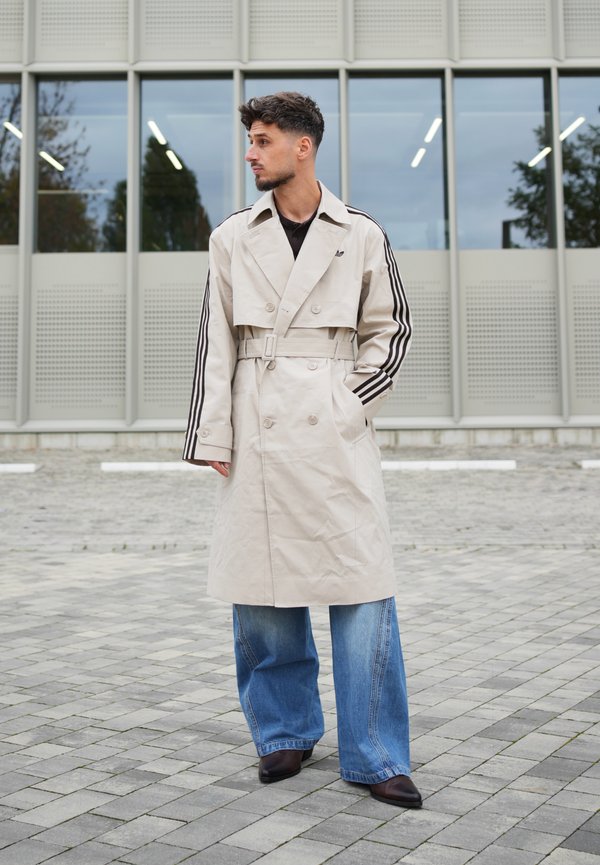 Un homme vêtu d’un trench beige ceinturé avec des bandes noires sur les manches, d’un jean bleu ample et de chaussures foncées se tient devant un bâtiment moderne.
