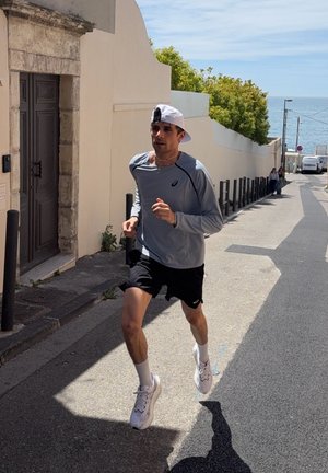 Hombre trotando por una calle iluminada por el sol, vistiendo una camiseta gris de manga larga, pantalones cortos negros, gorra blanca al revés y zapatillas blancas de correr.