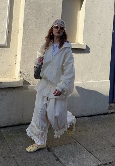 Mujer con chaqueta blanca de gran tamaño, pantalones blancos holgados con encaje, zapatos amarillos, sombrero beige de crochet, gafas de sol grandes y llevando un bolso gris en una acera.