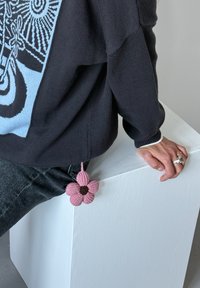 Pull noir en tricot avec un motif graphique bleu, associé à un jean foncé. Un pendentif en forme de fleur rose au crochet pend de la taille.