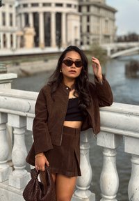 Chaqueta corta de ante marrón y falda a juego, camiseta negra debajo, complementada con gafas de sol oscuras y un bolso de mano de mimbre marrón.