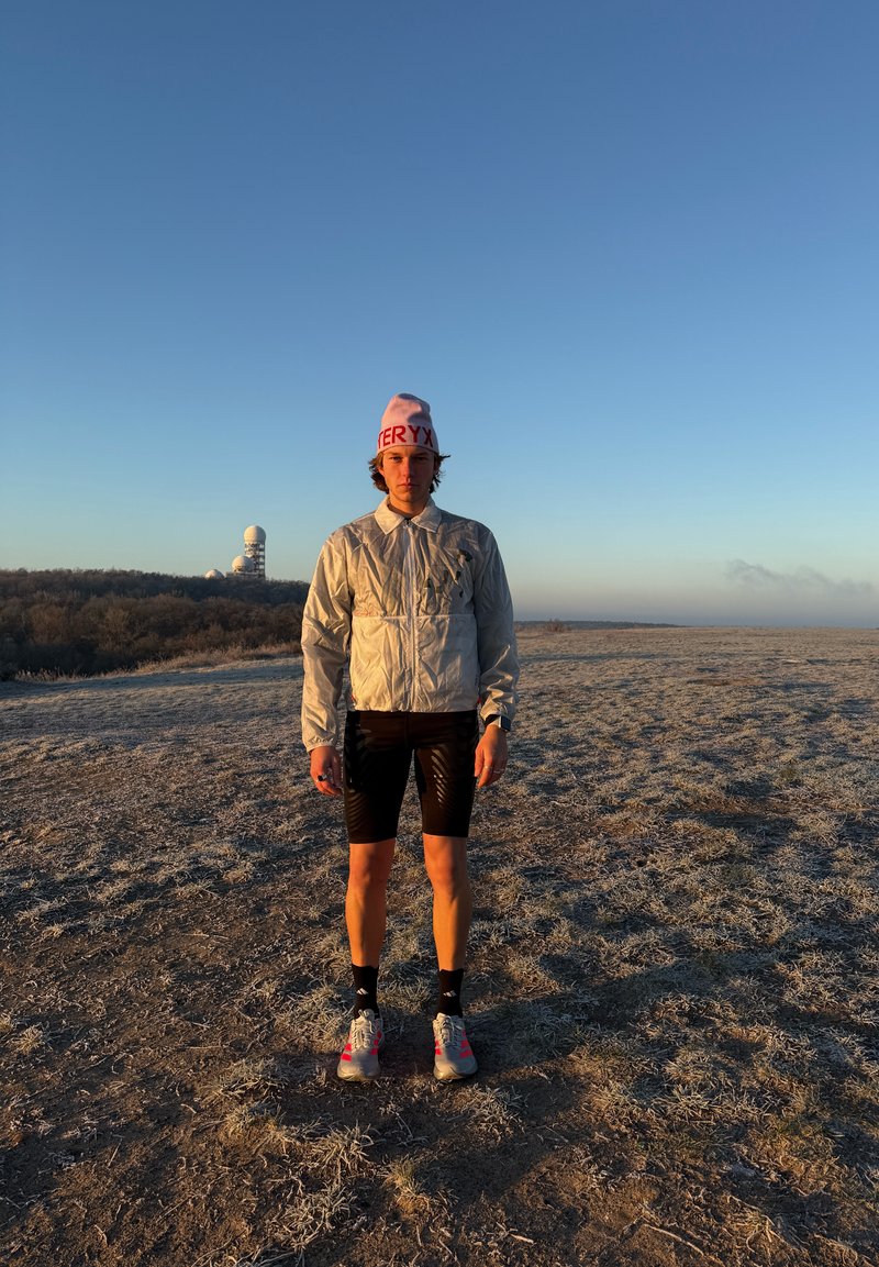 Ung person i refleksjakke, svarte shorts og rosa hatt som står på et frostdekket jorde ved soloppgang med radardomer på en fjern ås.