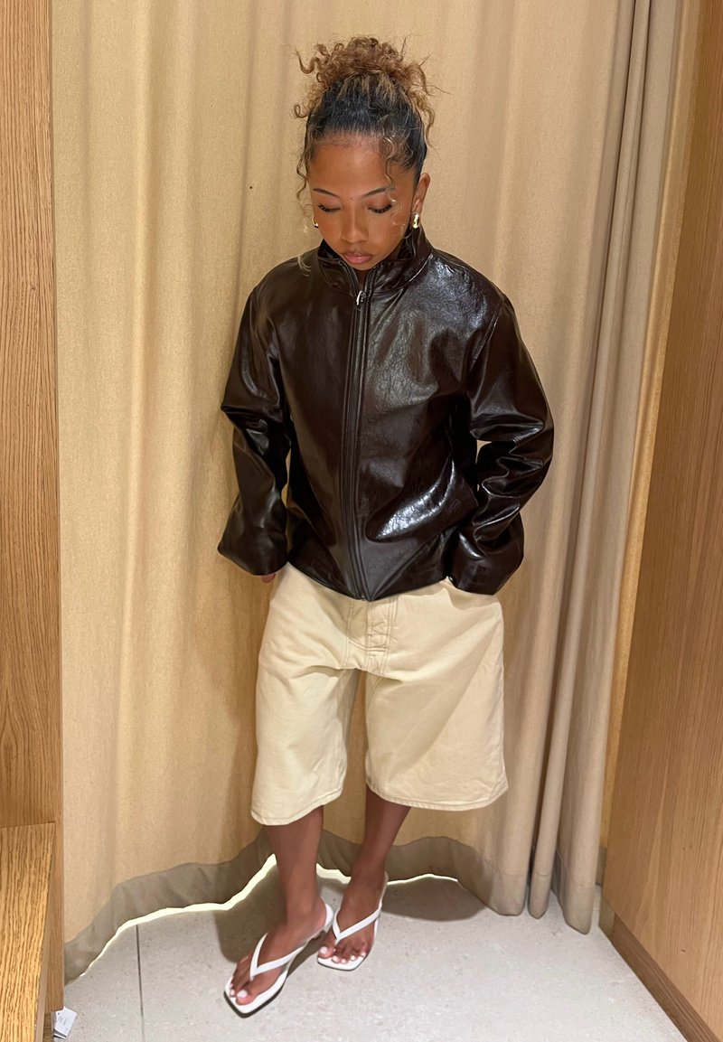 Veste en simili cuir marron avec fermeture éclair, coupe oversize ; associée à un short beige et des tongs blanches. Mise en scène devant un rideau neutre.