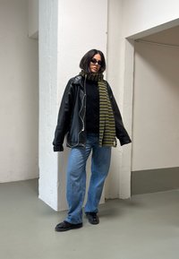 Kvinne iført svart lærjakke, stripete grønn skjerf, blå jeans, svarte sko og solbriller, stående innendørs mot hvite vegger.