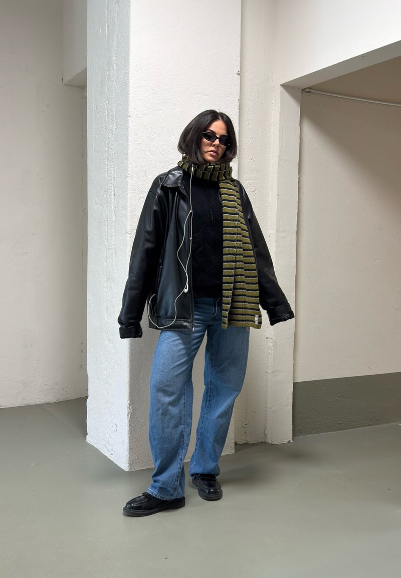 Vrouw in een zwart leren jas, gestreepte groene sjaal, blauwe jeans, zwarte schoenen en een zonnebril, staande binnen tegen witte muren.