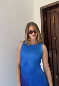 Mladá žena s rovnými plavými vlasy, která má na sobě sluneční brýle a modré šaty bez rukávů, stojí u tmavých dřevěných dveří.