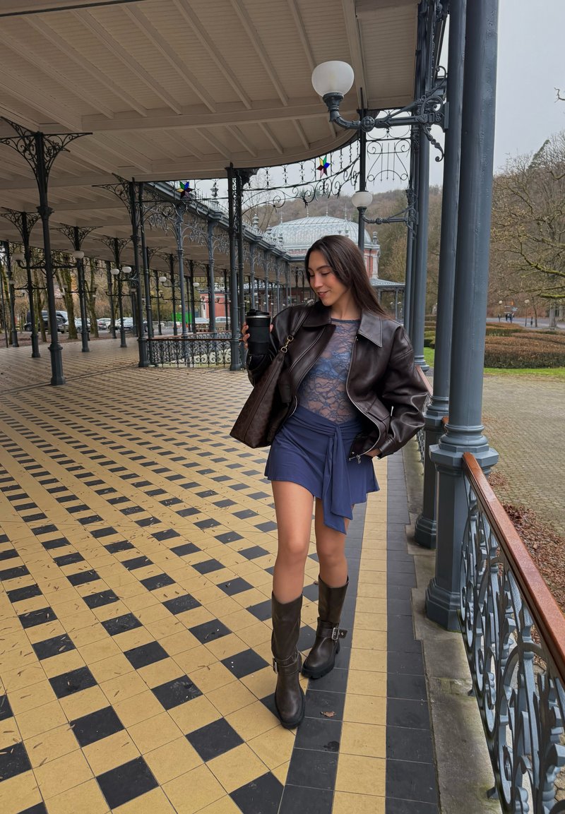Jeune femme marchant sur une terrasse carrelée à damier, portant une veste en cuir, une jupe bleue, des bottes, tenant une tasse à café de voyage noire.