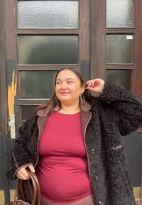 Musta kuvioitu takki, jossa on ruskeat nahkayksityiskohdat, päällä viininpunainen paita. Henkilöllä on ruskea laukku ja hän poseeraa lasioviseinän vieressä.