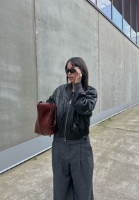 Svart läderbomberjacka, grå vida byxor, mörka solglasögon och en brun handväska. Släta texturer mot en betongbakgrund.