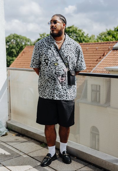 Camisa de manga corta en gris claro con estampado de leopardo negro y gráfico de un león, combinada con pantalones cortos negros y mocasines negros sobre calcetines blancos.