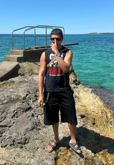 Camisa sin mangas negra con gráfico de la República de California, pantalones cortos negros, sandalias grises, de pie en una costa rocosa con agua turquesa de fondo.