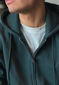 Tamsiai žalia su užtrauktuku striukė, su balta vidine apykakle, sidabriniu užtrauktuku ir raukšlėtomis rankovėmis. Užsidėta ant baltos t-shirt.