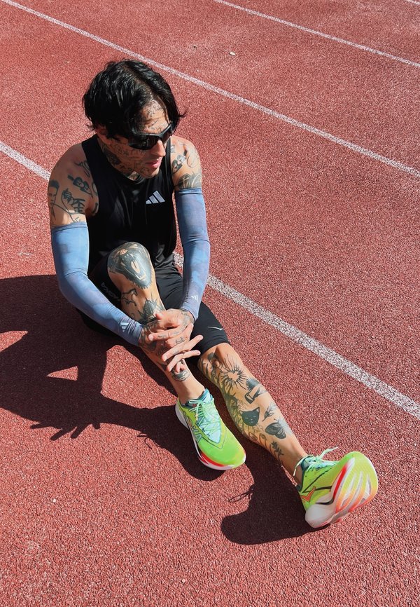 Getatoeëerde atleet met een zonnebril en neon hardloopschoenen zit op een rode baan en stretcht één been met beide handen in het zonlicht.