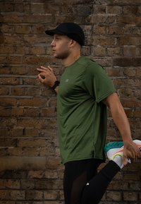 Idrettsutøver i en tettsittende grønn kortermet skjorte og svarte leggings, som strekker seg mot en murvegg, iført svart caps og fargerike løpesko.