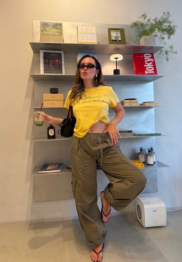 Una mujer con gafas de sol y una camisa amarilla posa con una bebida verde junto a unas estanterías con libros y objetos decorativos.