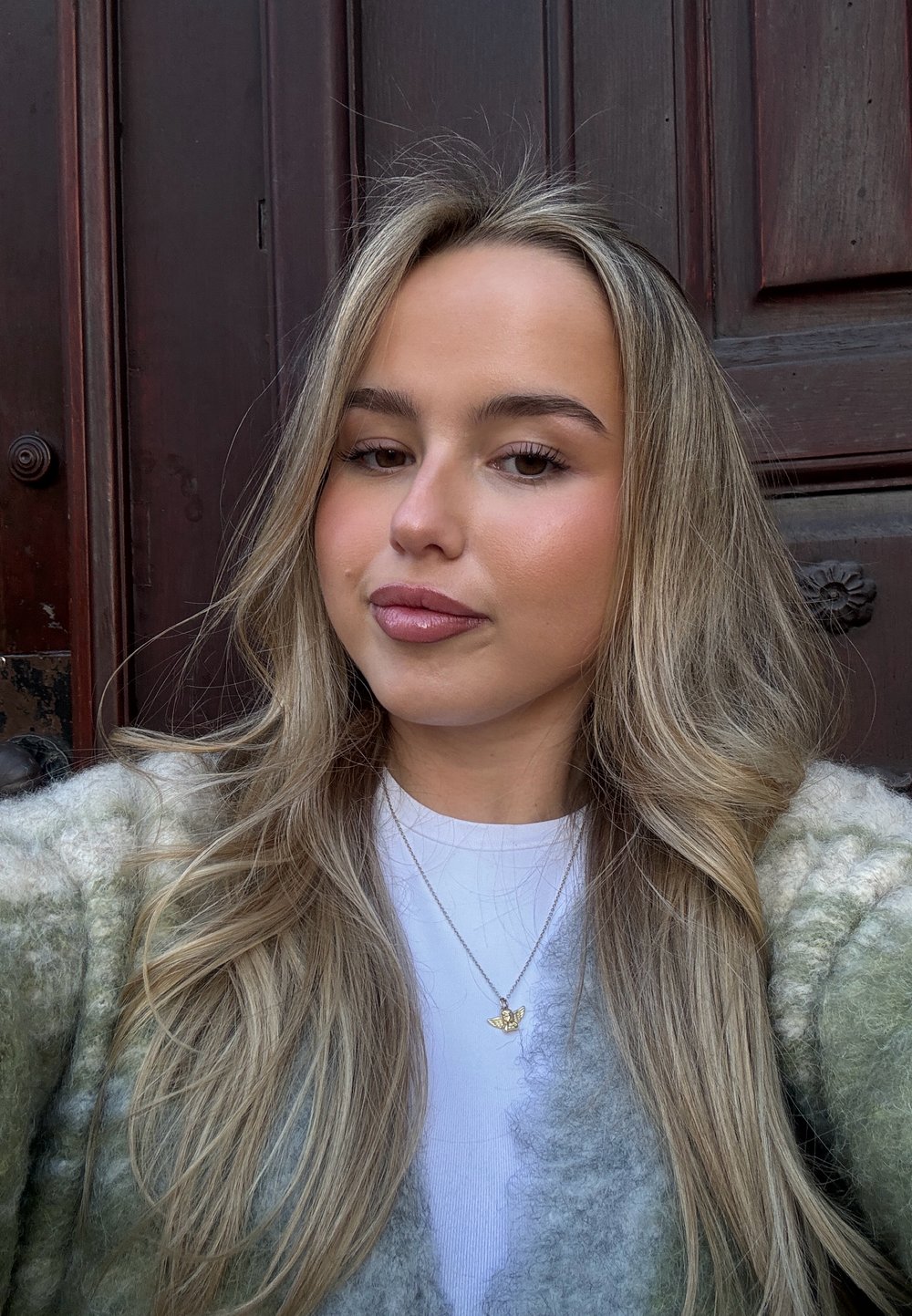 Φωτεινό πράσινο και κρεμ αφράτο πλεκτό κασκόλ πάνω από ένα λευκό πουκάμισο, με χρυσό κολιέ σε σχήμα πεταλούδας, και λείο, κυματιστό ξανθό μαλλί.