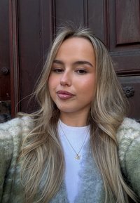 Φωτεινό πράσινο και κρεμ αφράτο πλεκτό κασκόλ πάνω από ένα λευκό πουκάμισο, με χρυσό κολιέ σε σχήμα πεταλούδας, και λείο, κυματιστό ξανθό μαλλί.