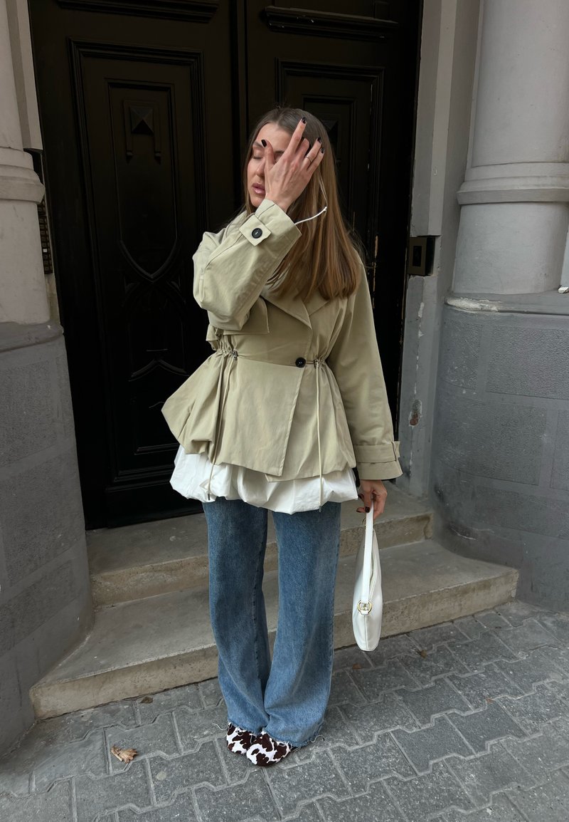 Femme aux longs cheveux portant une veste beige, un top blanc à couches, un jean bleu, des chaussures à motif vache, tenant un sac blanc, debout sur des marches près d'une porte sombre.