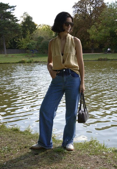 Top sin mangas beige, jeans de talle alto azul oscuro y zapatillas blancas. Lleva un pequeño bolso marrón cerca de un estanque.