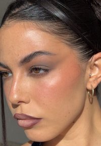 Mujer joven con ojos marrones y labios llenos, que lleva pendientes de aro dorados y pequeños pendientes de botón, cabello oscuro recogido con una diadema ancha.