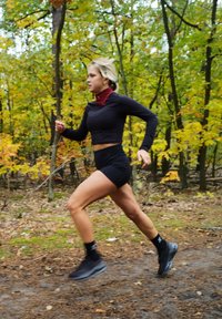 Juoda sportinė trumpa viršūnė ir šortai, bėgantys takeliu su rudens lapija. Užsidėję juodus batus ir raštuotas kojines.