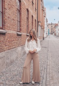 Fehér, hosszú ujjú blúz megkötő részlettel; bézs, bővérű nadrág; macskaköves utca, amelyet tégla és színes épületek szegélyeznek.