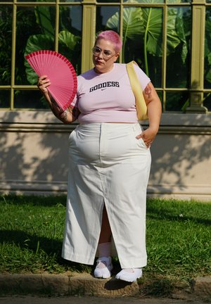 Camisa corta rosa con el texto "DIVA", falda maxi blanca y zapatos blancos. Sosteniendo un abanico rosa. Follaje verde de fondo.