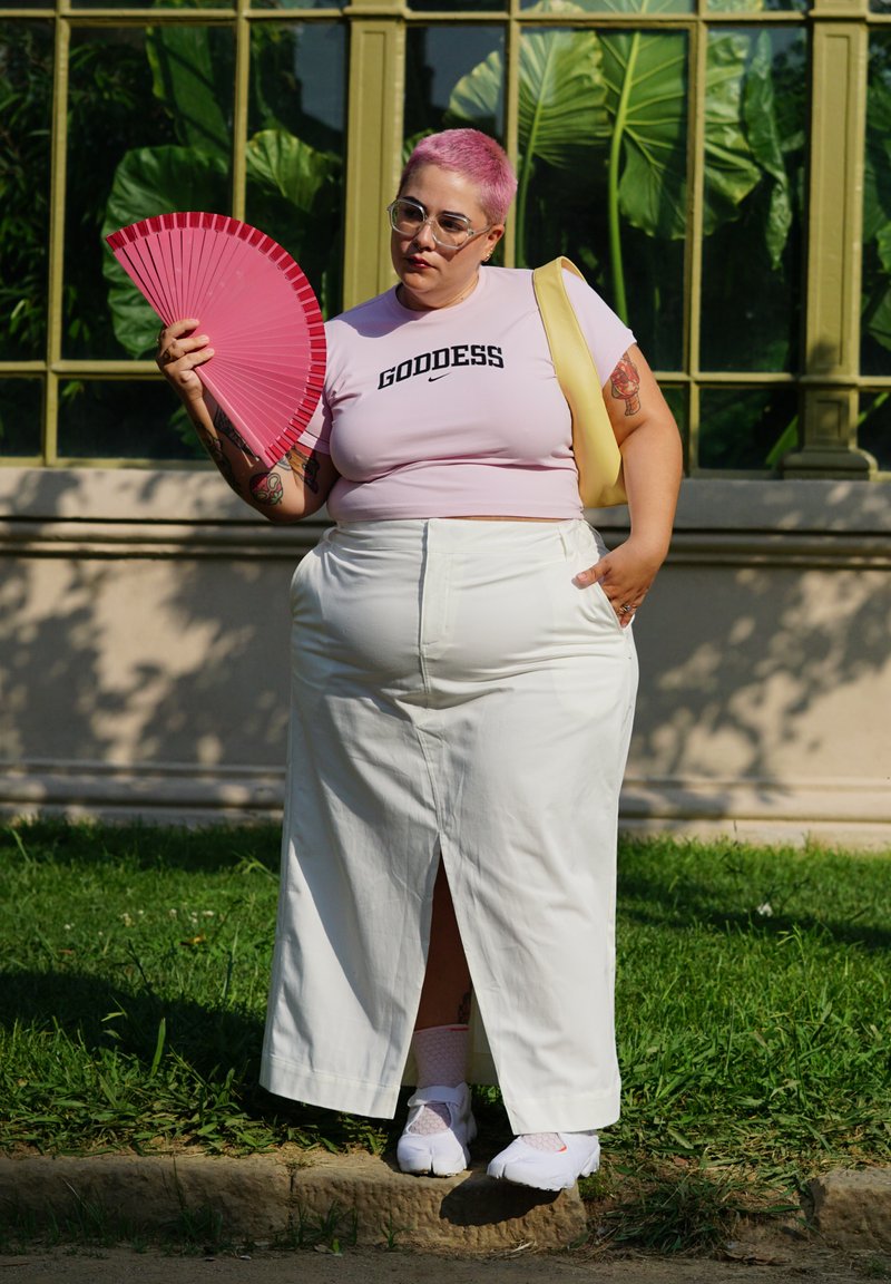 Chemise rose courte avec le texte "DÉESSE", jupe maxi blanche et chaussures blanches. Tenant un éventail rose. Fougères vertes en arrière-plan.
