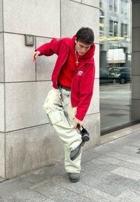 Κόκκινο φούτερ με φερμουάρ πάνω από κόκκινο μπλουζάκι με γραφικά, ελαφρώς φθαρμένα φαρδιά τζιν και σκούρες παντόφλες. Κρατώντας ένα μαύρο αντικείμενο, στέκεται απέναντι σε έναν γκρίζο τοίχο.