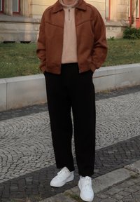 Brun ruskind zip-up jakke over en cremefarvet sweater, parret med sorte bukser og hvide sneakers. Står på mønstret brostenbelægning.