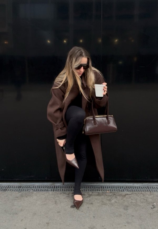 Kvinde i mørk frakke og solbriller justerer en brun spids sko, mens hun holder en hvid kop og en brun håndtaske mod en sort væg.