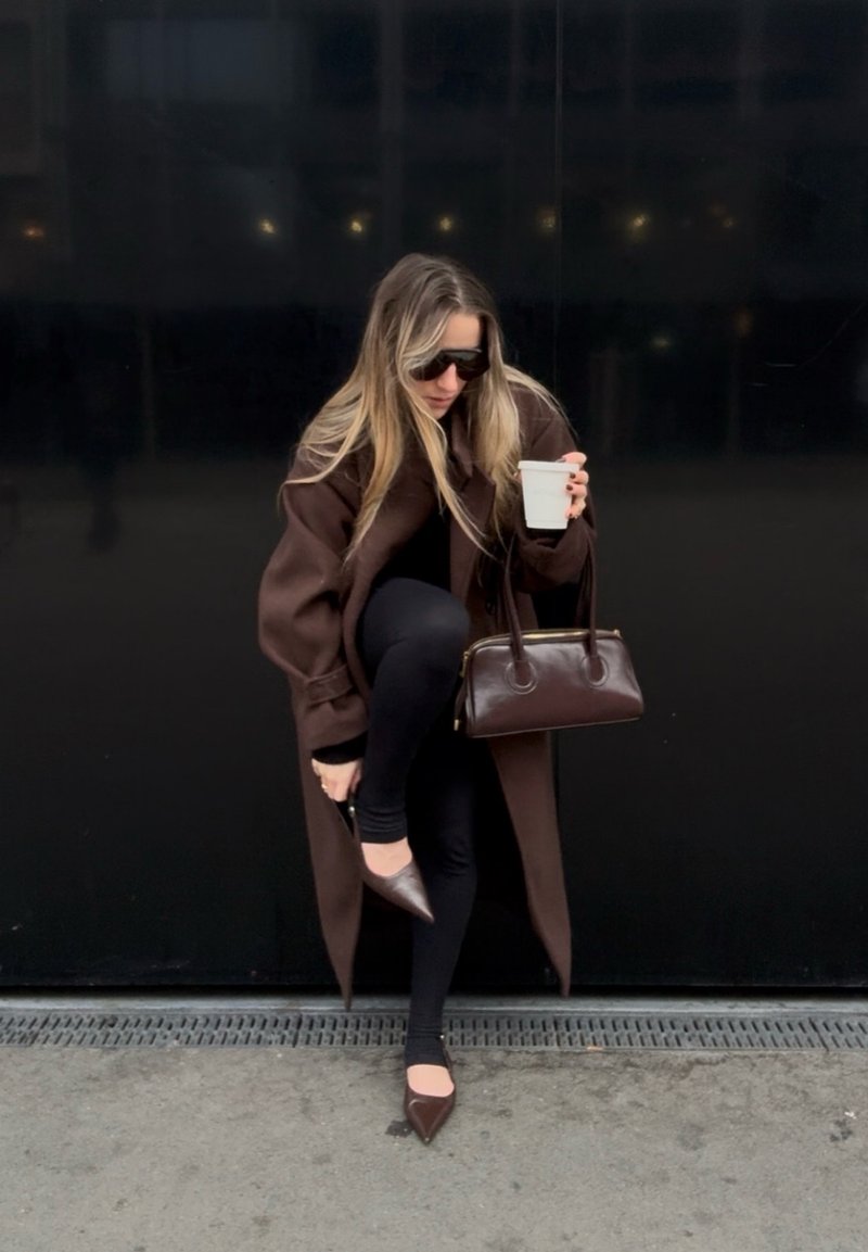 Γυναίκα με σκούρο παλτό και γυαλιά ηλίου ρυθμίζει καφέ μυτερό παπούτσι ενώ κρατάει ένα λευκό φλυτζάνι και μια καφέ τσάντα μπροστά σε μαύρο τοίχο.