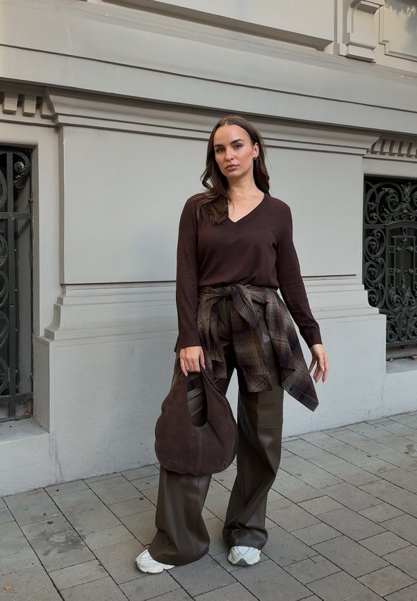 Mujer con camiseta de manga larga marrón oscuro, camisa de cuadros anudada a la cintura, pantalones de pierna ancha, zapatillas blancas, sosteniendo un bolso marrón, de pie en la acera.