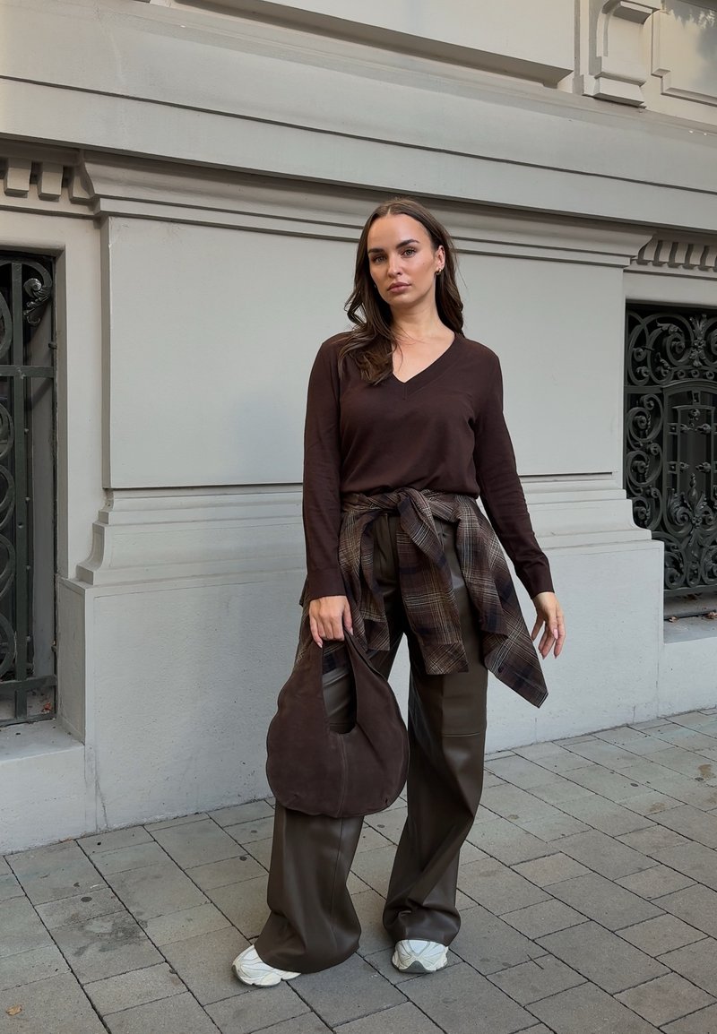 Femme portant un top à manches longues marron foncé, chemise à carreaux nouée à la taille, pantalon à jambes larges, baskets blanches, tenant un sac à main marron, debout sur le trottoir.