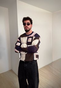 Jersey de punto con colores pastel y patrones cuadrados, gafas de sol negras y vaqueros oscuros, posando en un espacio interior minimalista.