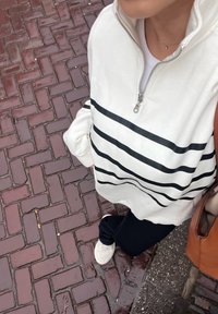 White zip-up sweatshirt with black horizontal stripes, teamed with dark trousers and light trainers. The background displays a brick pavement.