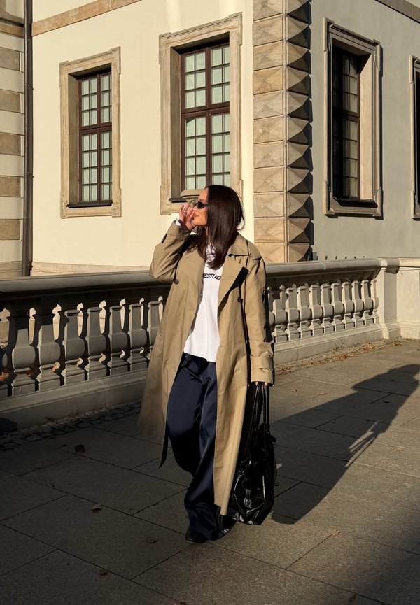 Mujer con gabardina beige, camisa blanca y gafas de sol caminando con bolso negro junto a un edificio histórico con grandes ventanas a la luz del sol.