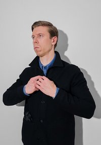 Manteau noir texturé avec boutons et col montant, superposé sur une chemise en jean bleu. Les poches présentent des accents noirs visibles.