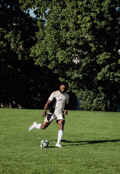 Camiseta deportiva en crema con acentos negros, combinada con pantalones cortos negros. Jugador pateando un balón de fútbol con patrones sobre hierba verde bajo la luz del sol.