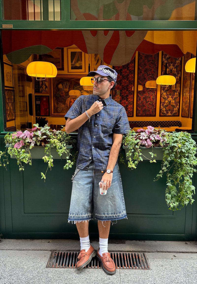 Chemise à manches courtes à carreaux bleus, short en denim avec bords effilochés, chaussettes blanches, mocassins marron. Plantes vertes et œuvres d'art en arrière-plan.