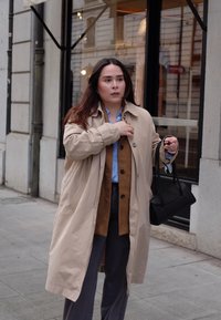 Beige trenchcoat over een bruin vest, lichtblauw shirt en grijze broek. Zwarte handtas met dunne schouderbanden, staande op een stadsstoep.