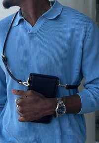 Svart lær crossover-veske sikret med et strop; brukt med en lys blå, ribbestrikket genser. Sølvklokke synlig på håndleddet.