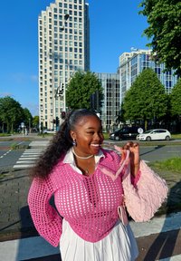 Rozā atvērta adījuma džemperis, kas uzvilkts virs balta krekla, kopā ar rozā frēzu somu. Apkārtnē redzami mūsdienīgi ēku kompleksi un koki.