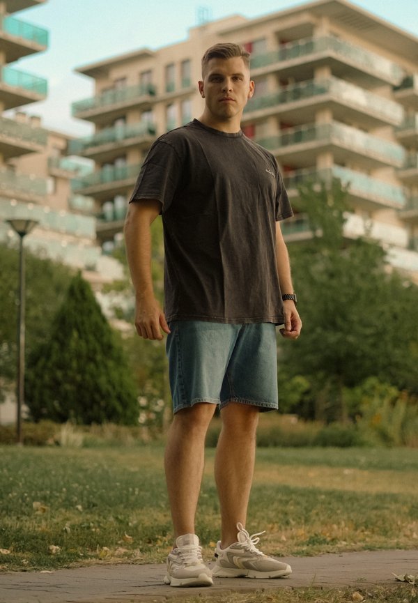 Νέος άνδρας με μαύρο μπλουζάκι, τζιν σορτς και λευκά αθλητικά παπούτσια, στέκεται σε ένα μονοπάτι με σύγχρονα διαμερίσματα και πράσινο στο φόντο.