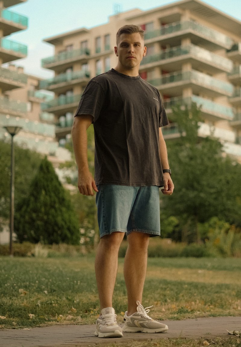 Jeune homme en t-shirt noir, short en jean et baskets blanches se tenant sur un chemin avec des immeubles modernes et de la verdure en arrière-plan.
